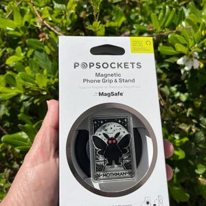 Puede incluir: Una caja blanca con un soporte de tel&eacute;fono magn&eacute;tico negro y plateado. El soporte presenta un dise&ntilde;o de mothman negro con ojos rojos. La caja dice "Magnetic Phone Grip & Stand" y "for MagSafe".