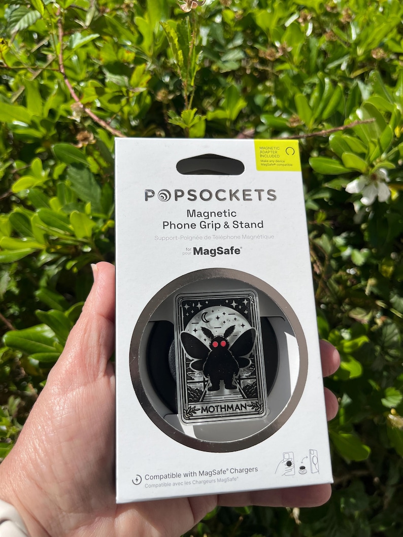 Puede incluir: Una caja blanca con un soporte de tel&eacute;fono magn&eacute;tico negro y plateado. El soporte presenta un dise&ntilde;o de mothman negro con ojos rojos. La caja dice "Magnetic Phone Grip & Stand" y "for MagSafe".
