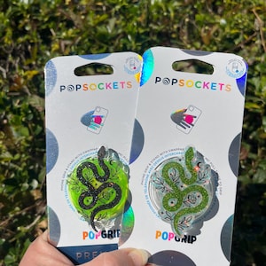 May include: Two PopSockets phone grips with a green snake design. The snake is outlined in black and has glitter accents. The background is a green and white floral pattern. The PopSockets are packaged in clear plastic packaging with the PopSockets logo and the words "PopGrip" printed on them.