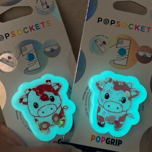 May include: Two PopSockets phone grips featuring a cartoon cow design with red and white accents, and a strawberry. The grips glow with a teal light. The packaging reads "POPSOCKETS" and "POPGRIP".