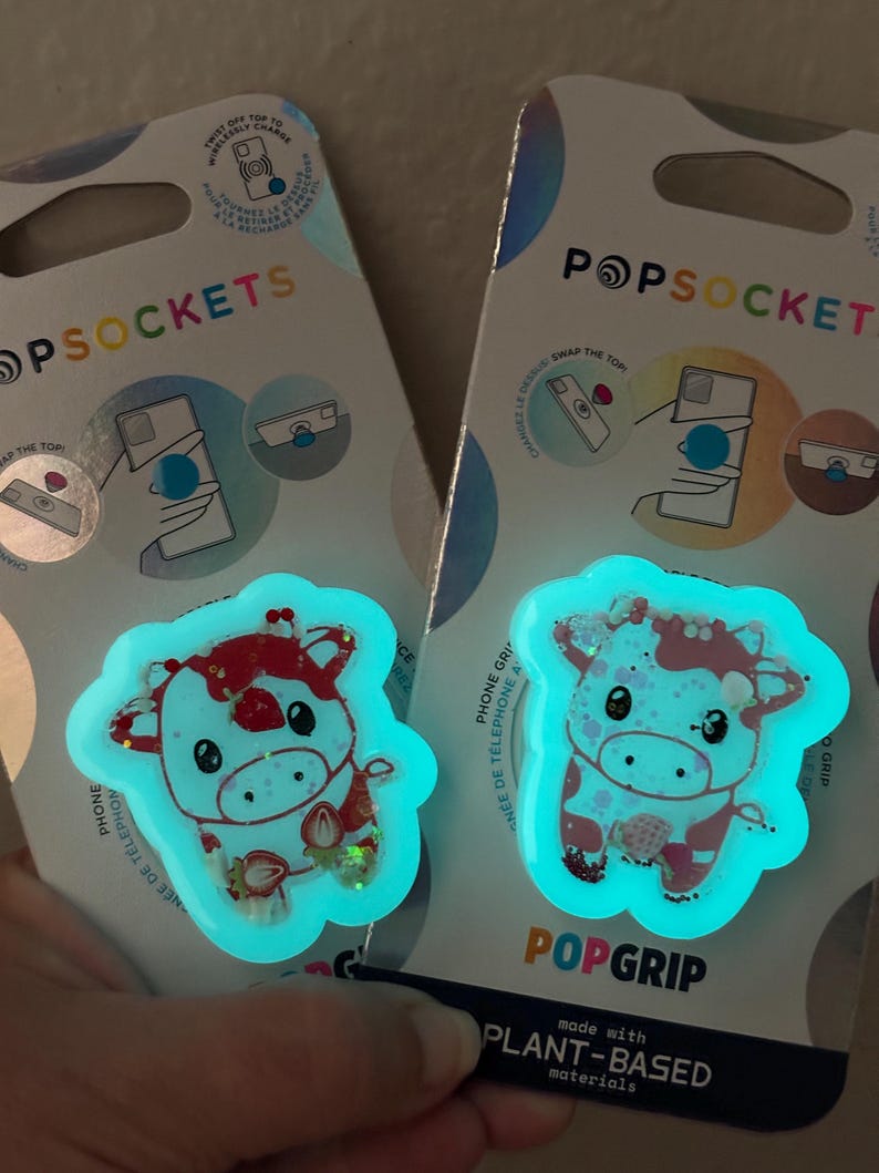 May include: Two PopSockets phone grips featuring a cartoon cow design with red and white accents, and a strawberry. The grips glow with a teal light. The packaging reads "POPSOCKETS" and "POPGRIP".