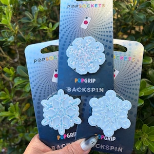 May include: Three PopSockets PopGrip Backspin phone grips with snowflake designs. The grips are white with iridescent glitter accents and are displayed on blue and silver packaging. The PopSockets logo is visible on the packaging.
