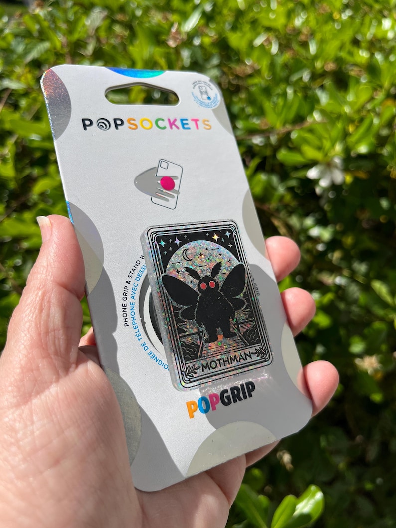 Puede incluir: Un agarre para tel&eacute;fono plateado e iridiscente con un dise&ntilde;o de mothman negro. El agarre tiene un acabado hologr&aacute;fico y el texto "Mothman" est&aacute; impreso en la parte inferior. El agarre es un producto de la marca PopGrip.