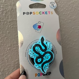 May include: A blue PopGrip phone grip with a black snake design and glitter accents. The snake is curled around a rose and leaves. The text "PopGrip" is printed on the packaging.