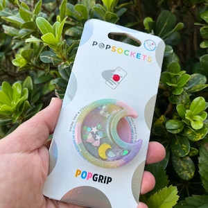 May include: A pastel pink, yellow, and blue PopSocket phone grip with a crescent moon, stars, and clouds. The PopSocket is on a white card with the text "PopSockets" and "PopGrip".
