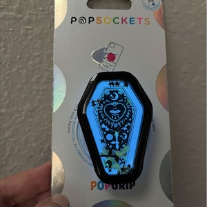 May include: A black and blue phone grip shaped like a coffin with a spooky eye, moon, and stars design. The grip is packaged on a white and silver background with the text "POPSOCKETS" and "POPGRIP".