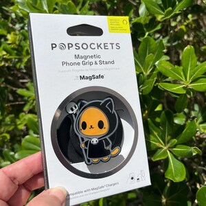 May include: A PopSockets Magnetic Phone Grip & Stand for MagSafe, featuring a black and orange cartoon cat design with a skull. The packaging is white with black text and includes a magnetic adapter.