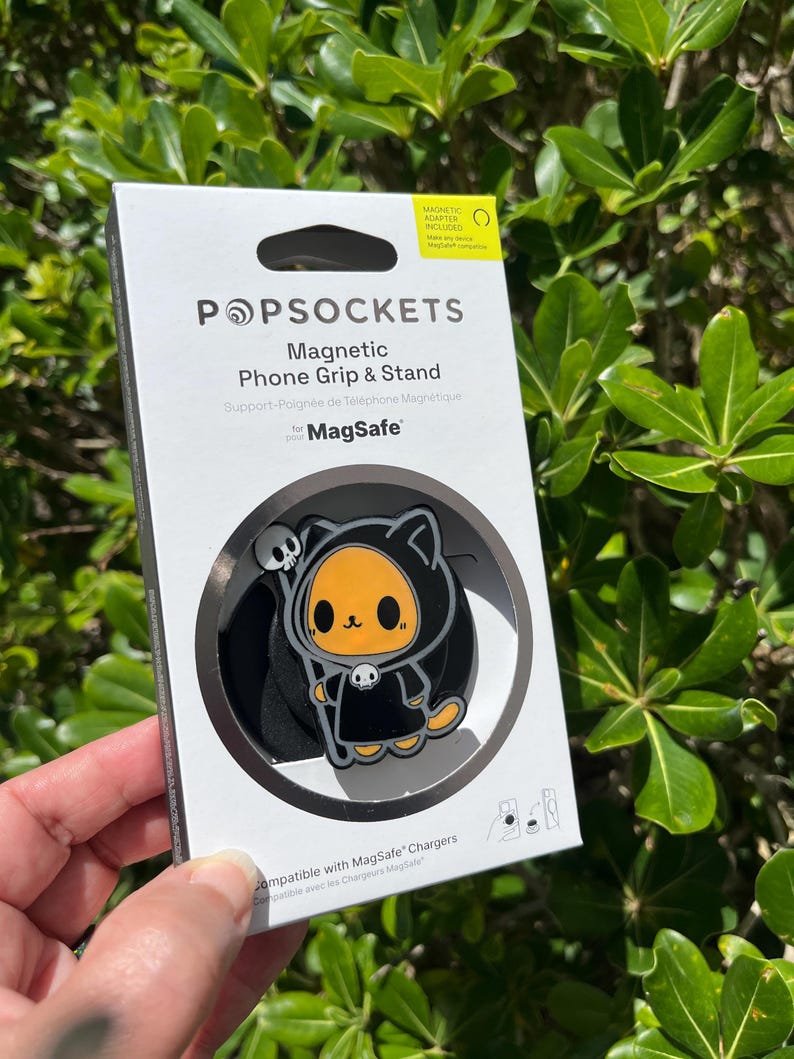 May include: A PopSockets Magnetic Phone Grip & Stand for MagSafe, featuring a black and orange cartoon cat design with a skull. The packaging is white with black text and includes a magnetic adapter.