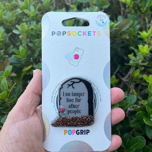 May include: A white and black phone grip with a tombstone design. The tombstone has a rose and glitter on it. The text on the tombstone reads "I no longer live for other people".