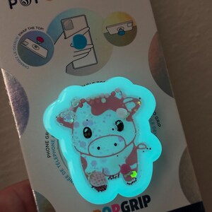 May include: A PopSockets PopGrip featuring a cartoon cow design with pink and white spots, outlined in a glowing blue. The PopGrip is attached to a white and blue backing with the PopSockets logo and instructions.