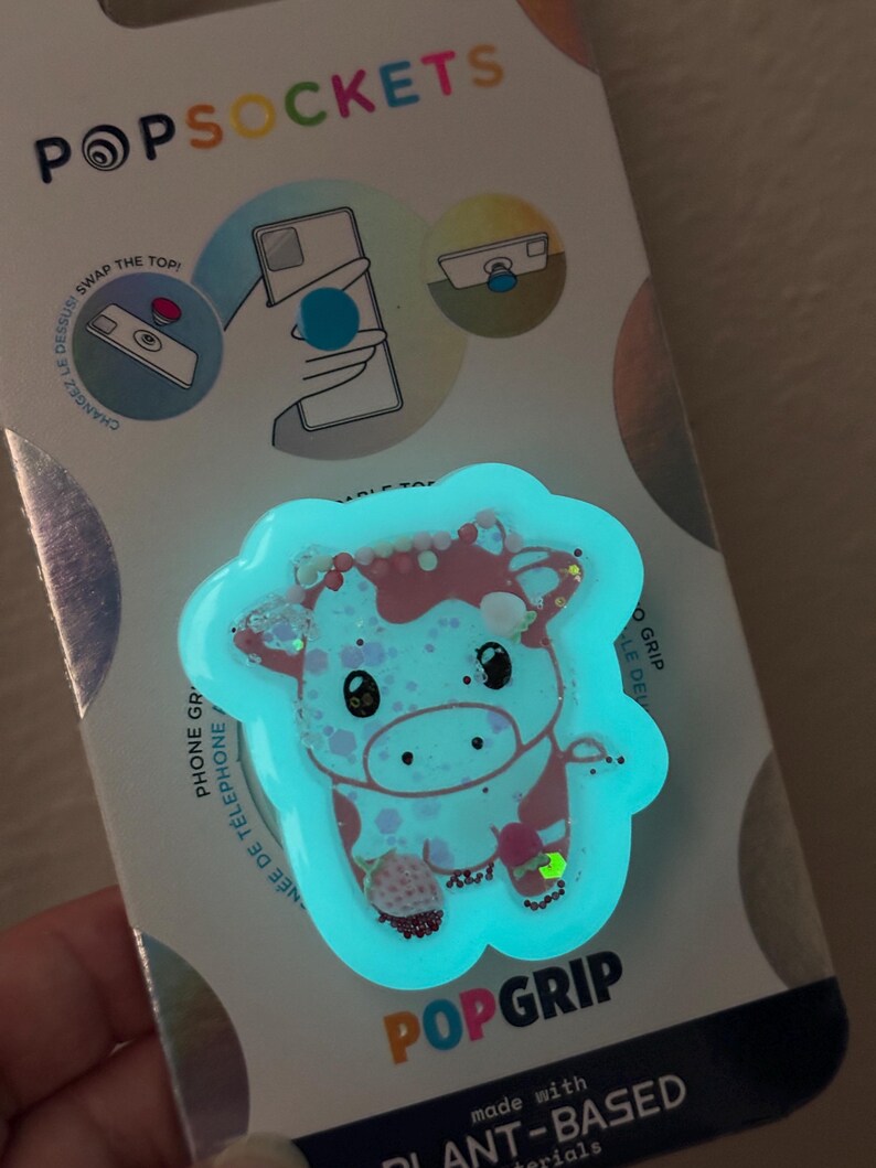 May include: A PopSockets PopGrip featuring a cartoon cow design with pink and white spots, outlined in a glowing blue. The PopGrip is attached to a white and blue backing with the PopSockets logo and instructions.