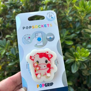 May include: A PopSocket phone grip featuring a red and white cow design with strawberry accents. The PopSocket is made with plant-based materials and has the PopSockets logo on the packaging.
