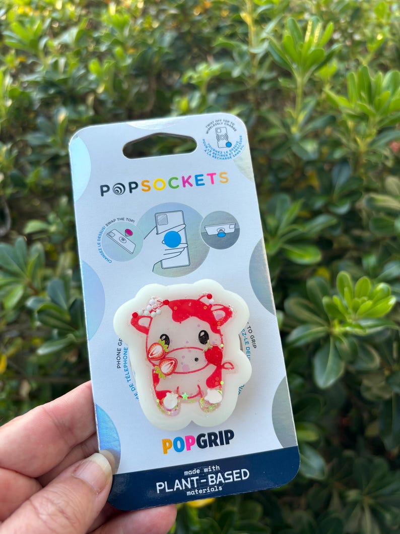 May include: A PopSocket phone grip featuring a red and white cow design with strawberry accents. The PopSocket is made with plant-based materials and has the PopSockets logo on the packaging.