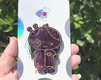 Puede incluir: Un agarre para teléfono PopSockets PopGrip con un diseño de muñeca cosida en morado y dorado. El PopGrip está sobre un respaldo blanco y plateado con el logotipo de PopSockets y las palabras "PHONE GRIP" y "POPGRIP".