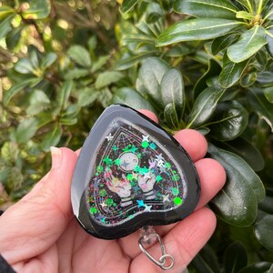 May include: Black heart-shaped resin charm with a glittery, iridescent design. The charm features a white ghost with outstretched hands and a black and white web design. The charm is attached to a clear plastic retractable badge holder.