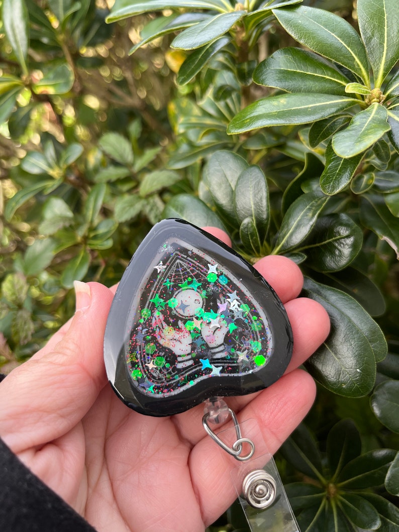 May include: Black heart-shaped resin charm with a glittery, iridescent design. The charm features a white ghost with outstretched hands and a black and white web design. The charm is attached to a clear plastic retractable badge holder.