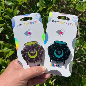 May include: Two PopSockets PopGrip phone accessories. One is a purple cauldron design with a green liquid, crescent moon, and stars. The other is a black cauldron with a teal liquid, crescent moon, and stars. The PopSockets packaging is white with the brand name.
