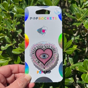 May include: A PopSockets PopGrip with a heart-shaped design featuring an eye and the text "KEEP YOUR BAD VIBES SHIELDED". The PopGrip is on a white and iridescent backing.