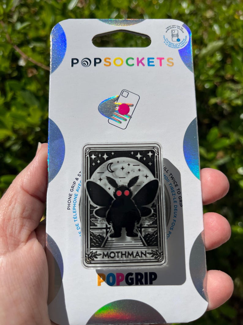 Puede incluir: Una agarradera para tel&eacute;fono de color plateado y negro con un gr&aacute;fico de Mothman, una criatura m&iacute;tica del folclore de Virginia Occidental. La agarradera tiene el texto "MOTHMAN" y "POPGRIP" impreso en ella. El empaque tambi&eacute;n incluye el texto "FILL TWICE TO GRIP" y "MOI! TIREZ-LE DEUX FOIS PO-".