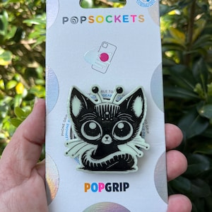 May include: A black and white PopGrip phone grip with a cartoon cat design. The cat has large eyes and a white tail. The PopGrip is attached to a white card with the words "PopSockets" and "PopGrip" printed on it.