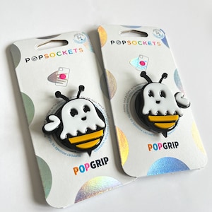May include: A white ghost with black eyes and a yellow and black striped body, wearing a black bee headband, is attached to a PopGrip phone stand. The PopGrip is on a white and iridescent background with the words "PopSockets" and "PopGrip" printed on it.