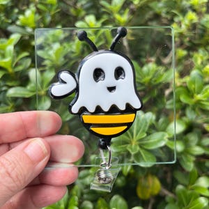 May include: A badge reel featuring a white ghost-shaped cover over a black and yellow striped bee body. The character has two antennae, a smiling face, and one raised arm. The design is mounted on a clear square acrylic plate with a metal clip.