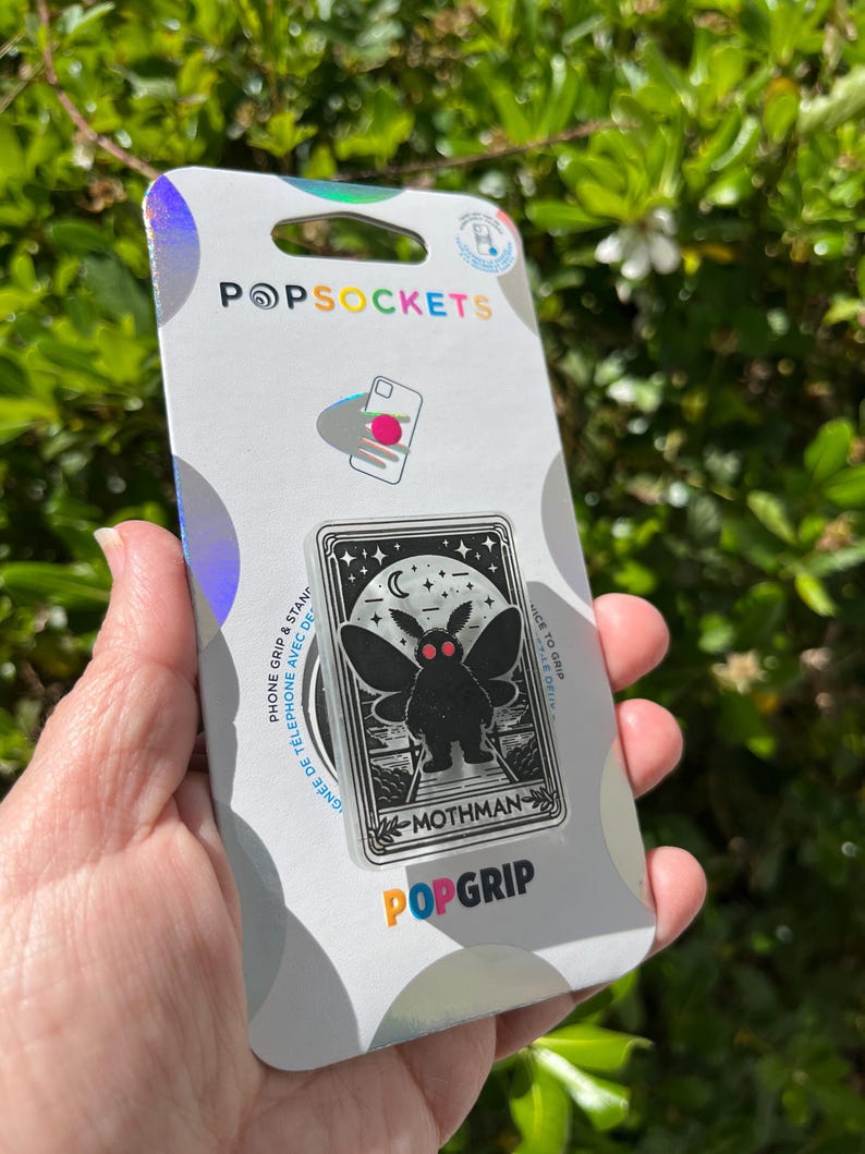 Puede incluir: Una agarradera para tel&eacute;fono PopSockets plateada con una ilustraci&oacute;n en blanco y negro de Mothman, una criatura cr&iacute;ptida con grandes ojos rojos y alas. El texto "Mothman" est&aacute; impreso debajo de la ilustraci&oacute;n. El texto "PopGrip" est&aacute; impreso en colores arco&iacute;ris debajo de la ilustraci&oacute;n.