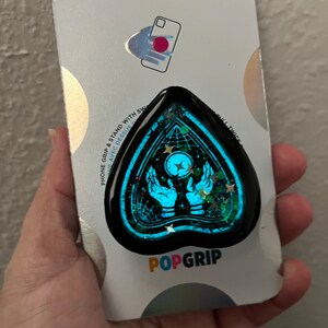 Pu&ograve; includere: Un PopGrip per telefono blu e nero con un design glitterato che presenta un paio di mani che tengono una luna crescente con stelle. Il testo "POPGRIP" &egrave; stampato sulla confezione.