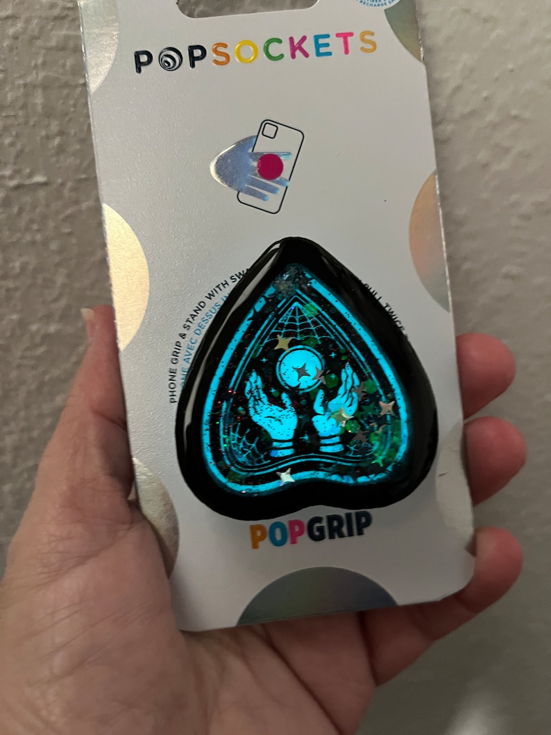 Pu&ograve; includere: Un PopGrip per telefono blu e nero con un design glitterato che presenta un paio di mani che tengono una luna crescente con stelle. Il testo "POPGRIP" &egrave; stampato sulla confezione.