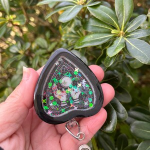 May include: Black heart-shaped resin keychain with a clear background and glitter. The keychain features a black and white image of a hand holding a crystal ball with stars and a web design. The keychain is attached to a silver retractable badge holder.