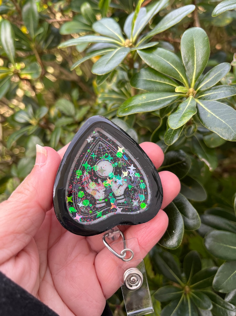 May include: Black heart-shaped resin keychain with a clear background and glitter. The keychain features a black and white image of a hand holding a crystal ball with stars and a web design. The keychain is attached to a silver retractable badge holder.