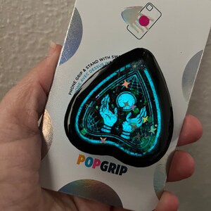 Pu&ograve; includere: Un'impugnatura per telefono nera con un design blu e verde scintillante che presenta una mano con una sfera di cristallo e stelle. L'impugnatura ha la forma di una lacrima e ha il testo "POPGRIP" stampato su di essa.