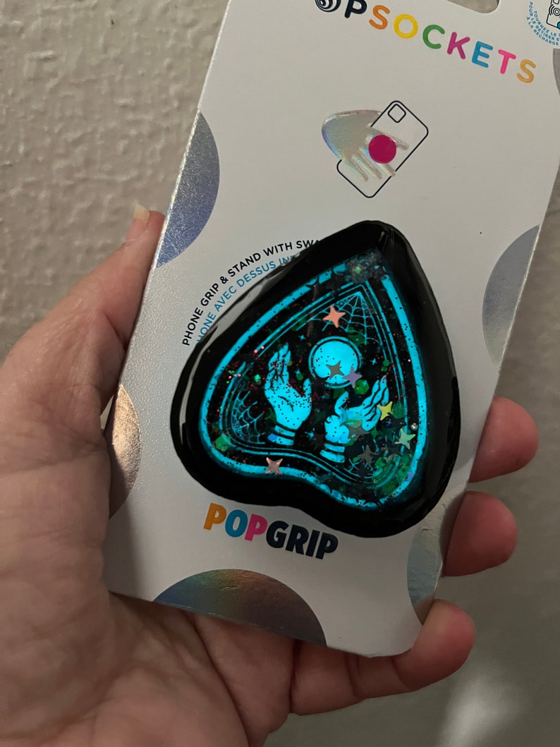 Pu&ograve; includere: Un'impugnatura per telefono nera con un design blu e verde scintillante che presenta una mano con una sfera di cristallo e stelle. L'impugnatura ha la forma di una lacrima e ha il testo "POPGRIP" stampato su di essa.