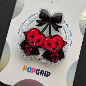 May include: A glittery acrylic phone grip featuring two red heart-shaped vampire characters with black capes and fangs, topped with a black bow. The accessory is mounted on a white backing card with the text POPGRIP printed in orange and black.