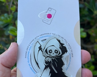 May include: A white PopGrip phone grip with a black and white illustration of the Grim Reaper holding a scythe. The text "PopGrip" is printed in colorful letters below the image. The text "Phone Grip & Stand with Swappable Top. Try Me! Pull Twice to Grip. -Le Deux Fois Poi-  Poignée de Téléphone Avec" is printed around the image.