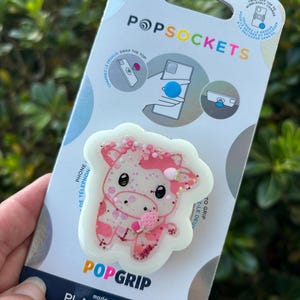 May include: A PopSockets PopGrip featuring a pink and white cow design with glitter accents. The PopGrip is made with plant-based materials and is attached to a white and blue backing with the PopSockets logo.