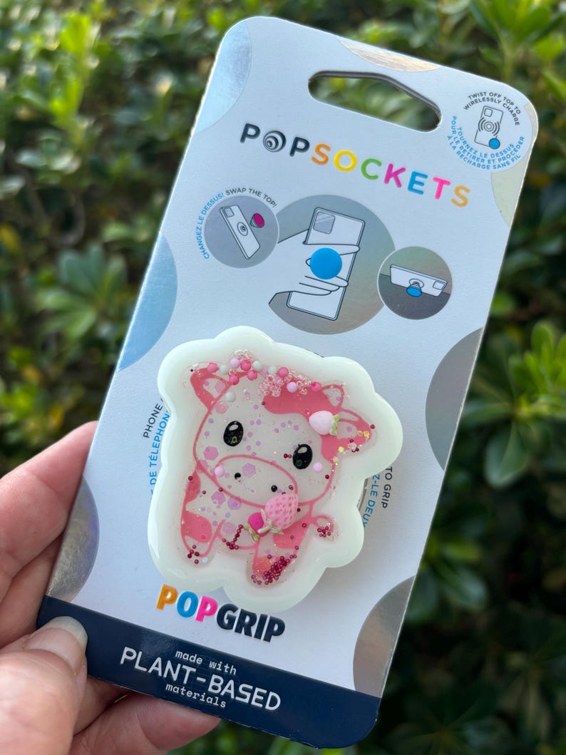 May include: A PopSockets PopGrip featuring a pink and white cow design with glitter accents. The PopGrip is made with plant-based materials and is attached to a white and blue backing with the PopSockets logo.