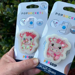 May include: Two PopSockets phone grips. Each grip features a cartoon cow design with pink and red accents, glitter, and a white border. The packaging reads "POPSOCKETS" and "POPGRIP."