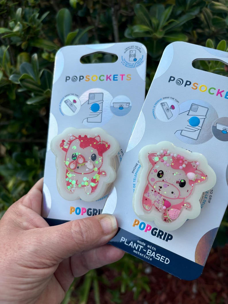 May include: Two PopSockets phone grips. Each grip features a cartoon cow design with pink and red accents, glitter, and a white border. The packaging reads "POPSOCKETS" and "POPGRIP."