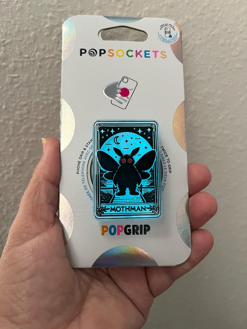 Puede incluir: Un pin de esmalte azul y negro con un mothman con ojos rojos. El pin est&aacute; en una tarjeta blanca con el texto "PopGrip" y "Mothman" impreso en &eacute;l.