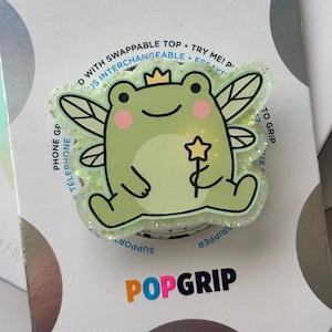 May include: A light green frog-shaped phone grip featuring a fairy frog with wings, a small crown, and a star-tipped wand. The design includes pink cheeks and glitter accents. The packaging text reads POPGRIP and includes instructions.