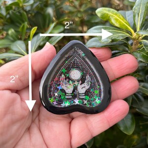 Pu&ograve; includere: Un ciondolo in resina nero a forma di cuore con un design bianco di due mani che tengono una luna e stelle. Il ciondolo &egrave; di circa 5 cm di diametro e ha una finitura scintillante.