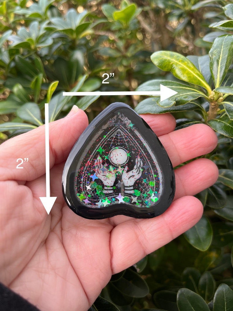 Pu&ograve; includere: Un ciondolo in resina nero a forma di cuore con un design bianco di due mani che tengono una luna e stelle. Il ciondolo &egrave; di circa 5 cm di diametro e ha una finitura scintillante.