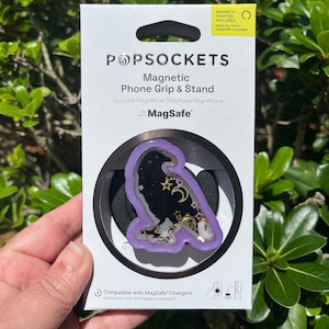 May include: A PopSockets magnetic phone grip and stand with a purple raven design. The raven is filled with gold stars, moons, and other decorative elements. The packaging includes the text "Magnetic Adapter Included" and "Compatible with MagSafe Chargers."