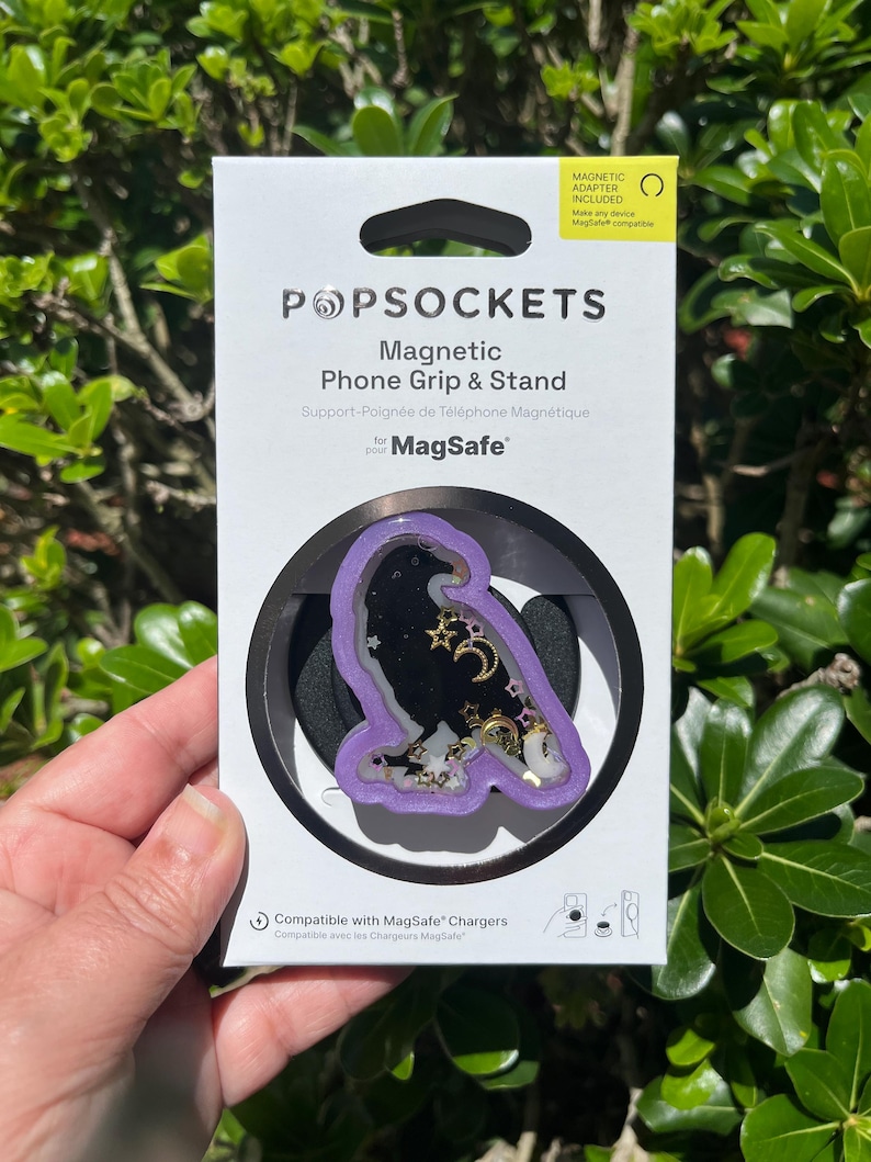May include: A PopSockets magnetic phone grip and stand with a purple raven design. The raven is filled with gold stars, moons, and other decorative elements. The packaging includes the text "Magnetic Adapter Included" and "Compatible with MagSafe Chargers."