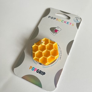 May include: A yellow honeycomb PopGrip phone stand with a small bee on top. The PopGrip is on a white background with the words "PopSockets" and "PopGrip" printed on the packaging.