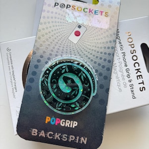 May include: A PopSockets PopGrip Backspin phone grip and stand. The grip features a swirling design with teal and silver accents. The packaging includes the PopSockets logo and text indicating compatibility with MagSafe chargers.