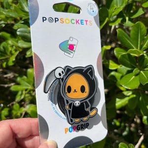 May include: A PopSockets PopGrip featuring a cartoon cat dressed as the Grim Reaper. The cat is orange and black, with a skull on its hood. The PopGrip is attached to a white and silver backing with the PopSockets logo.