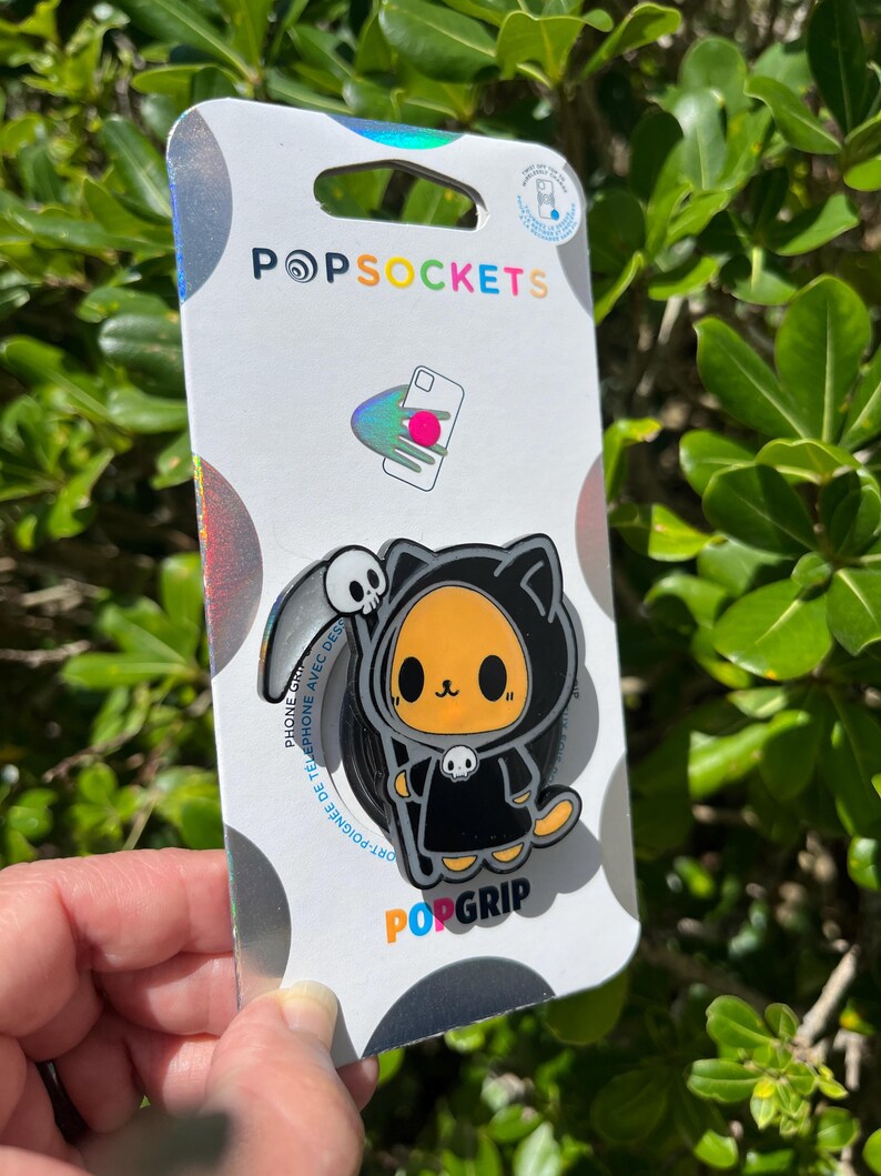 May include: A PopSockets PopGrip featuring a cartoon cat dressed as the Grim Reaper. The cat is orange and black, with a skull on its hood. The PopGrip is attached to a white and silver backing with the PopSockets logo.