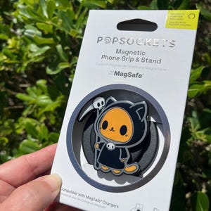 May include: A PopSockets magnetic phone grip and stand featuring a cartoon Grim Reaper cat design. The black and orange character holds a scythe and wears a hooded robe. The packaging includes the text "Magnetic Phone Grip & Stand" and "Compatible with MagSafe Chargers."
